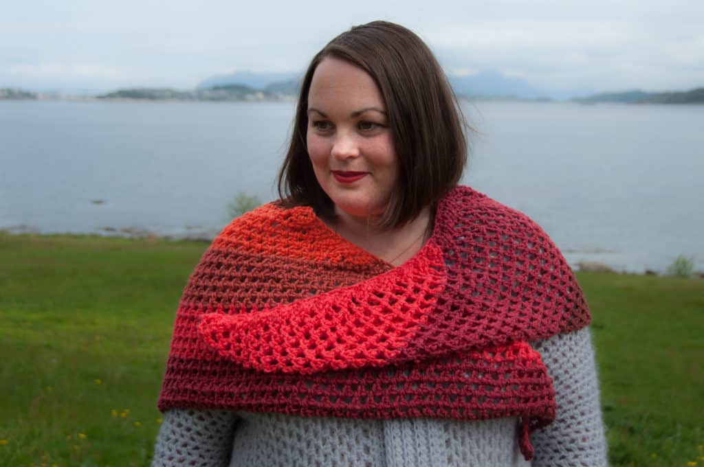 Tunisian crochet shawl in gradient shades of red and orange, draped over a person standing by a lake.