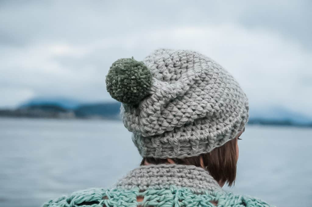 Gray crochet beanie with a green pom-pom, made with bulky yarn, worn by a person near a body of water.