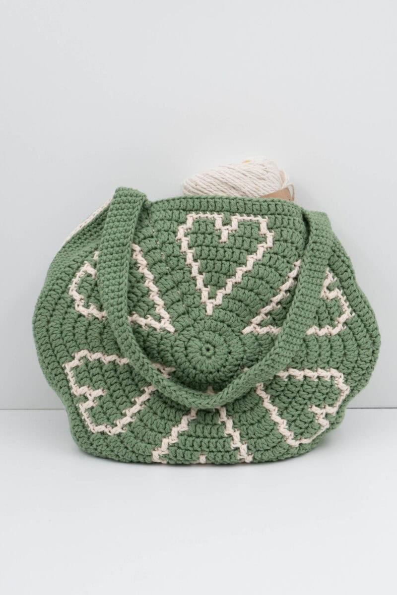Green crochet bag with heart patterns in cream, featuring round handles and a ball of cream yarn peeking out.
