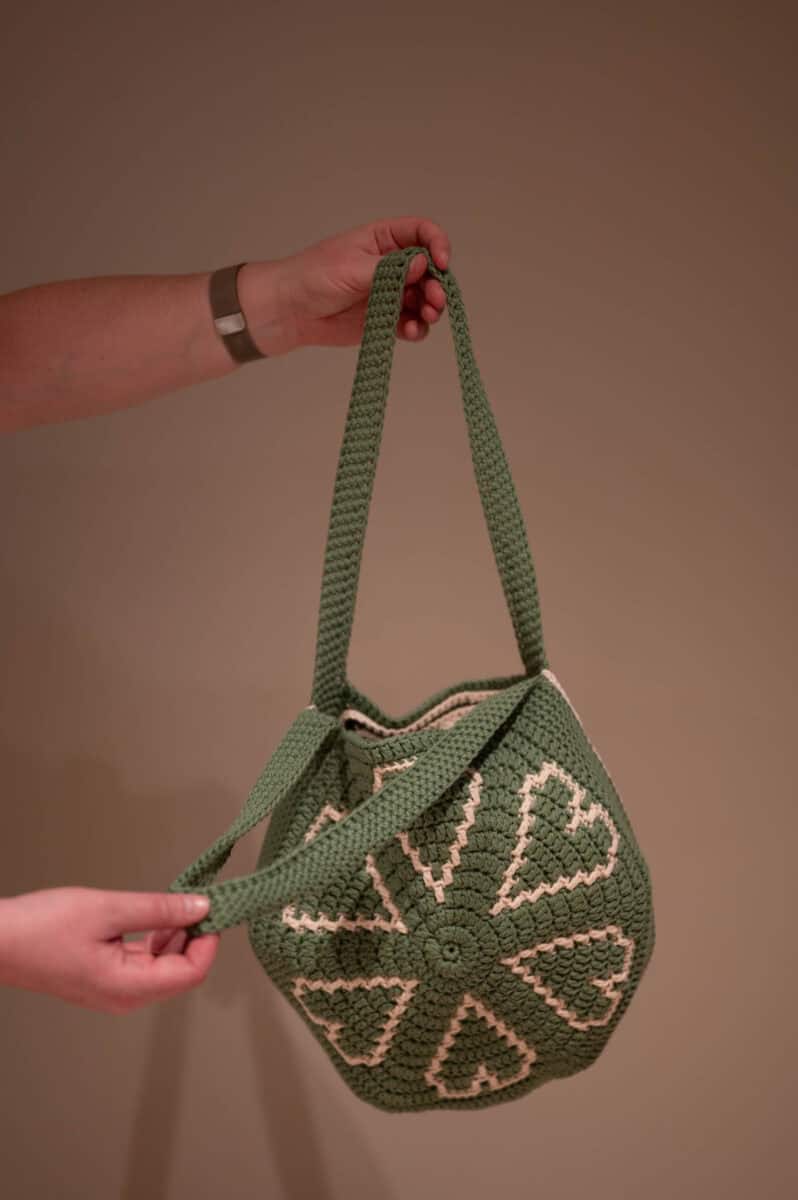 Green crochet bag with cream heart patterns, held by two hands against a plain background.